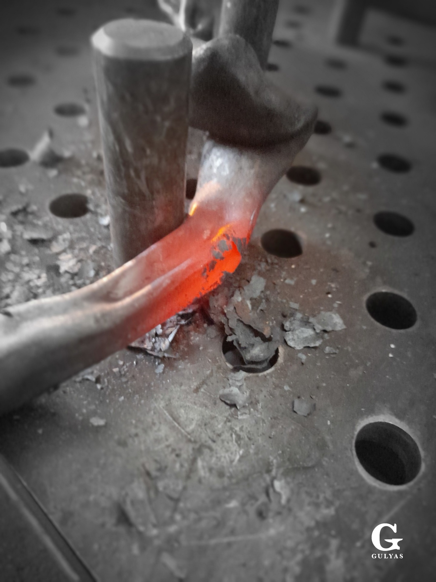 A detailed view of an orange, glowing hot piece of metal being pressed and bent between steel pegs on a blacksmith's swage block, with tiny scales flaking off the metal